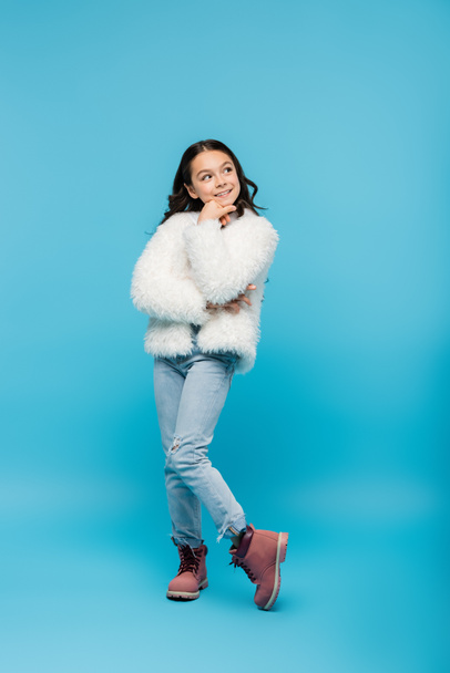 full length of cheerful preteen girl in faux fur jacket posing on blue - 写真・画像