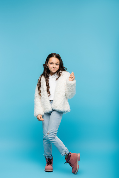 full length of positive preteen girl in faux fur jacket posing on blue - Φωτογραφία, εικόνα