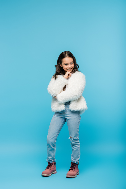 full length of positive preteen girl in faux fur jacket and winter boots posing on blue - Foto, Imagen