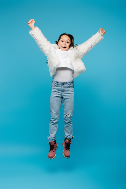 full length of positive preteen girl in faux fur jacket and winter boots jumping on blue - Foto, Imagem