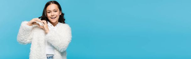 positive preteen girl in white faux fur jacket showing heart with hands isolated on blue, banner - Фото, зображення