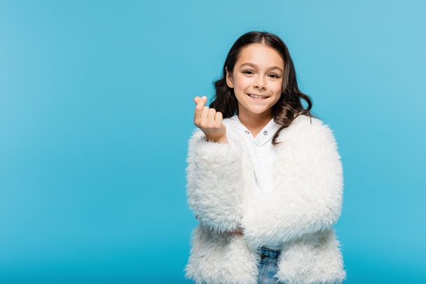 szczęśliwy nastolatek dziewczyna w biały faux futro kurtka patrząc na aparat i pokazując znak serca z palce izolowane na niebieski - Zdjęcie, obraz