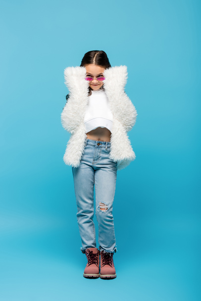 full length of smiling preteen girl in pink sunglasses and white faux fur jacket on blue - Fotó, kép