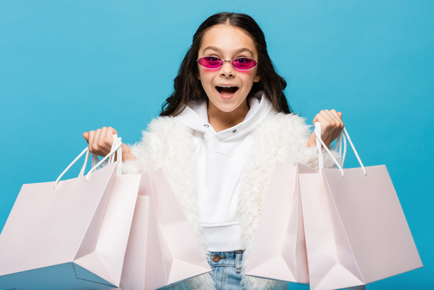 amazed preteen girl in pink sunglasses and faux fur jacket holding shopping bags isolated on blue - Photo, Image