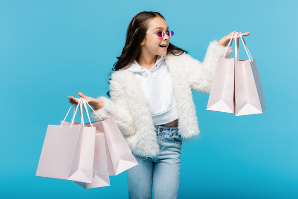 excited preteen girl in pink sunglasses and faux fur jacket holding shopping bags isolated on blue - Photo, Image