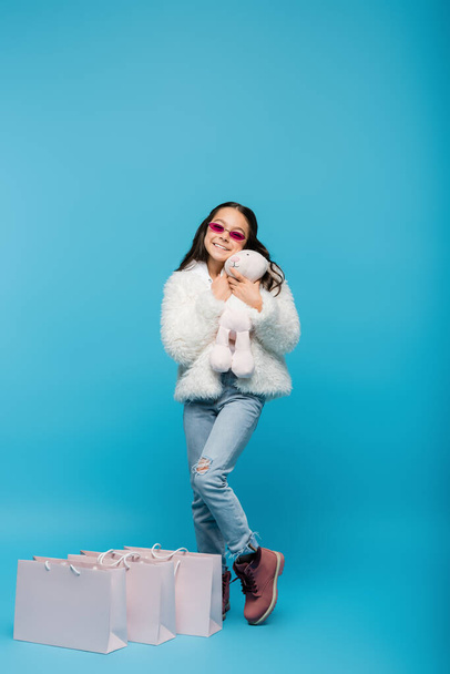 full length of happy preteen girl in pink sunglasses and faux fur jacket holding soft bunny near shopping bags on blue - Photo, Image