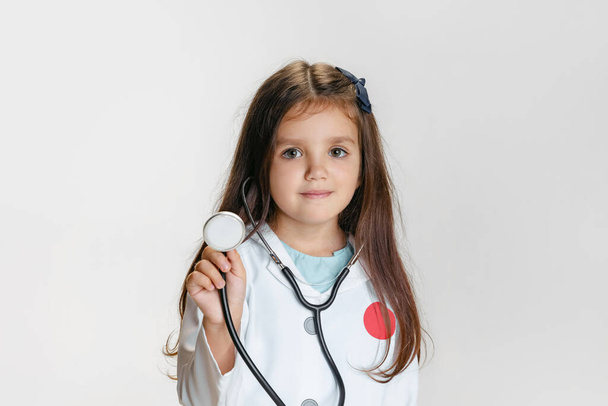 Close-up portrait of cute little girl, child in image of doctor wearing white lab coat with stethoscope isolated on white studio background - 写真・画像