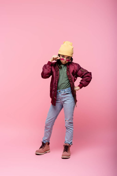полная длина стильная девочка-подросток в модном наряде регулируя солнцезащитные очки и позируя с рукой на бедре на розовый  - Фото, изображение