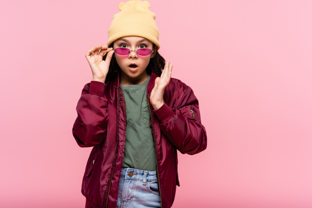 shocked preteen girl in trendy outfit adjusting sunglasses isolated on pink - Photo, Image
