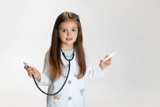 Close-up portrait of cute little girl, child in image of doctor wearing white lab coat with stethoscope isolated on white studio background - Photo, Image