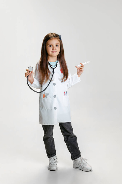 Portrait of cute beautiful girl, child in image of doctor wearing white lab coat posing isolated on white studio background - Foto, immagini