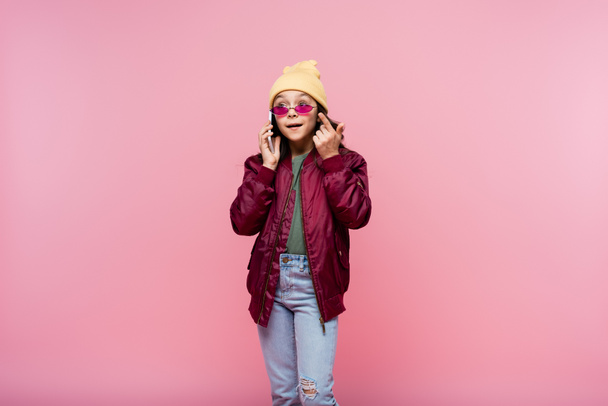 niña preadolescente en traje elegante y gafas de sol hablando en el teléfono inteligente aislado en rosa - Foto, imagen