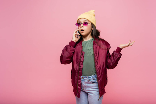wstrząśnięty nastolatek dziewczyna w stylowy strój i okulary przeciwsłoneczne rozmowy na smartfonie podczas gestykulując izolowane na różowy - Zdjęcie, obraz