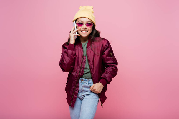 joyful preteen girl in stylish outfit and sunglasses talking on smartphone isolated on pink - Fotó, kép