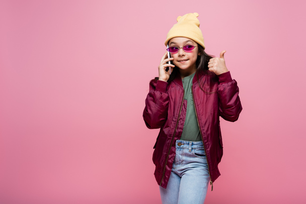 joyful preteen girl in stylish outfit and sunglasses talking on smartphone and showing thumb up isolated on pink - Photo, Image