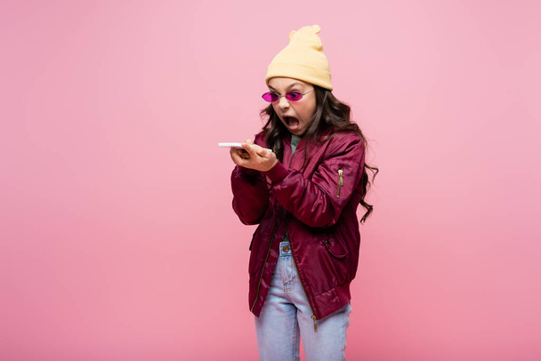 angry preteen girl in stylish outfit and sunglasses looking at smartphone and screaming isolated on pink - Foto, Imagen