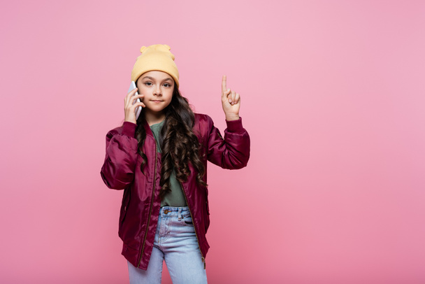 preteen kid in stylish outfit talking on cellphone and pointing with finger isolated on pink  - Zdjęcie, obraz
