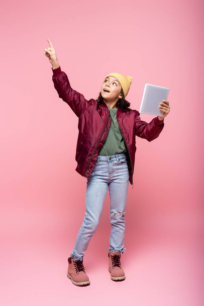 full length of stylish preteen girl holding digital tablet and pointing with finger on pink  - Fotó, kép