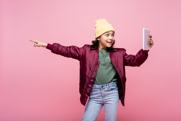 happy preteen girl in trendy outfit holding digital tablet and pointing with finger isolated on pink  - Photo, Image