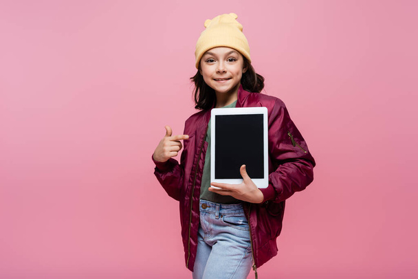 felice preteen girl in abito alla moda che punta con il dito al tablet digitale con schermo bianco isolato su rosa  - Foto, immagini