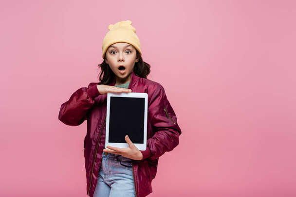 shocked preteen girl in trendy outfit holding digital tablet with blank screen isolated on pink  - 写真・画像