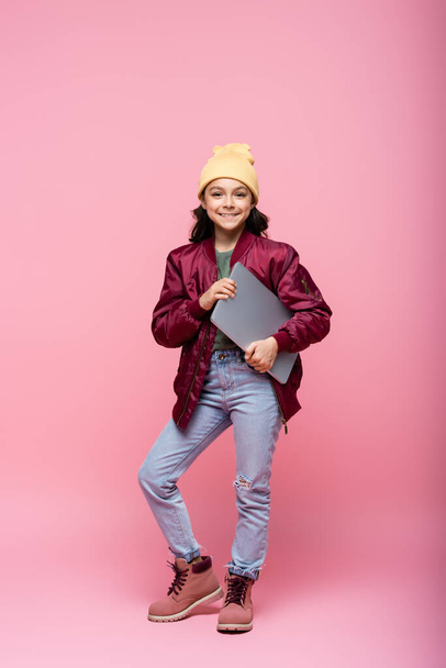 full length of stylish preteen girl in winter outfit posing with laptop and smiling on pink  - Foto, immagini