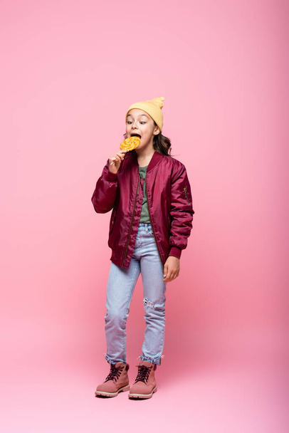 full length of preteen girl in stylish outfit eating lollipop on pink  - Фото, зображення