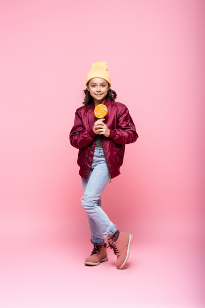 full length of cheerful preteen girl in winter outfit holding lollipop on pink  - Photo, Image