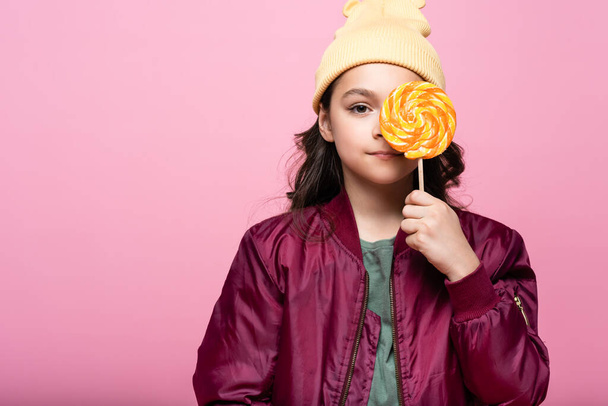 stylish preteen girl in winter outfit holding lollipop and covering eye isolated on pink  - Photo, Image