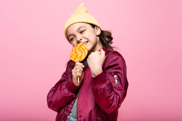 stylish preteen girl in winter outfit holding lollipop and smiling isolated on pink  - Fotoğraf, Görsel