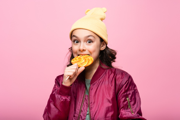 trendy preteen girl in winter outfit biting lollipop isolated on pink - Photo, Image