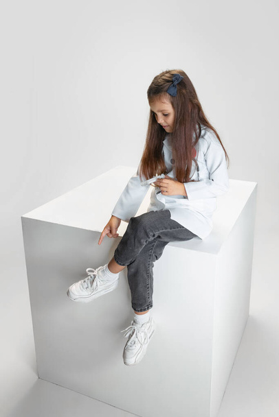 One cute little girl, child in image of doctor wearing white lab coat sitting on huge box isolated on white studio background - Photo, Image