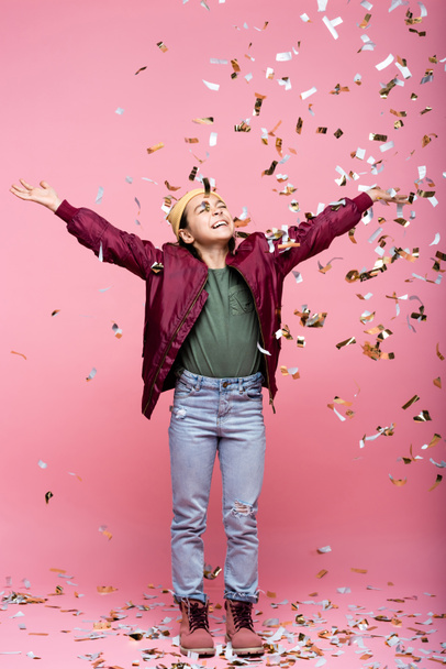 full length of joyful preteen girl in stylish outfit smiling near falling confetti on pink  - Zdjęcie, obraz