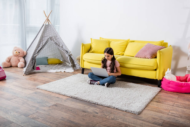niño preadolescente sentado en la alfombra y el uso de ordenador portátil en la sala de estar moderna  - Foto, imagen