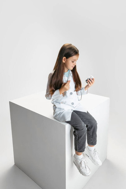 Jedna roztomilá holčička, dítě na obrázku doktora v bílém laboratorním plášti sedí na obrovské krabici izolované na bílém pozadí studia - Fotografie, Obrázek