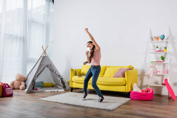 glückliches Mädchen mit drahtlosen Kopfhörern, das das Smartphone hält, während es im modernen Wohnzimmer tanzt  - Foto, Bild
