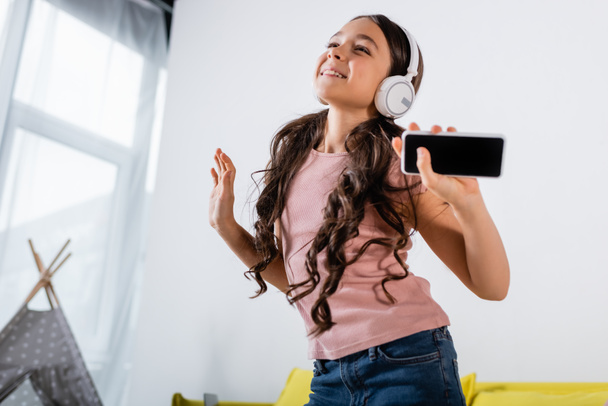 happy preteen girl in wireless headphones holding smartphone while dancing at home - Foto, Imagen