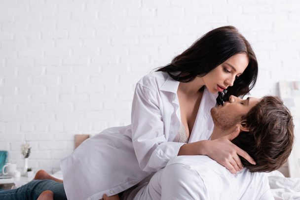 passionate woman in unbuttoned shirt embracing young man in bedroom - Photo, image