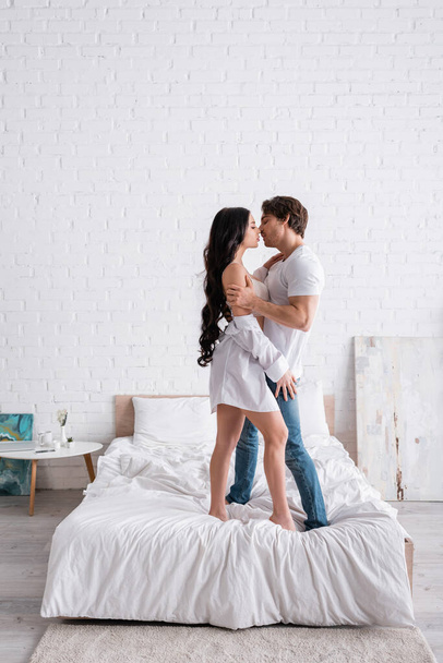 full length view of sexy couple embracing and kissing while standing on bed - Valokuva, kuva