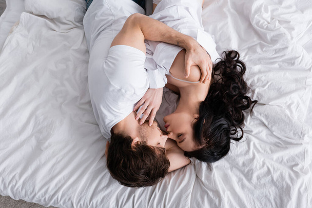 vista superior de pareja joven apasionada con los ojos cerrados abrazando en la ropa de cama blanca - Foto, Imagen