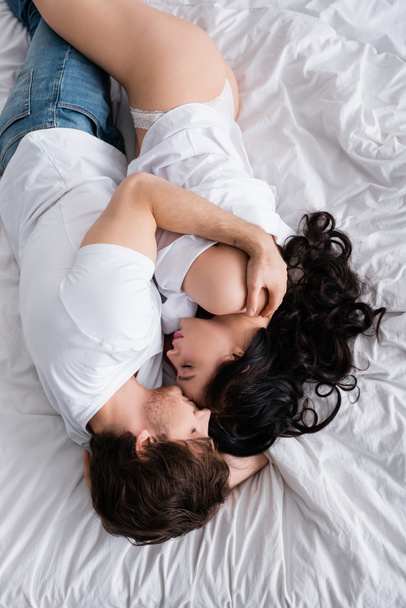 над головой вид мужчины в джинсах обнимающего сексуальную женщину в рубашке и трусиках на кровати - Фото, изображение