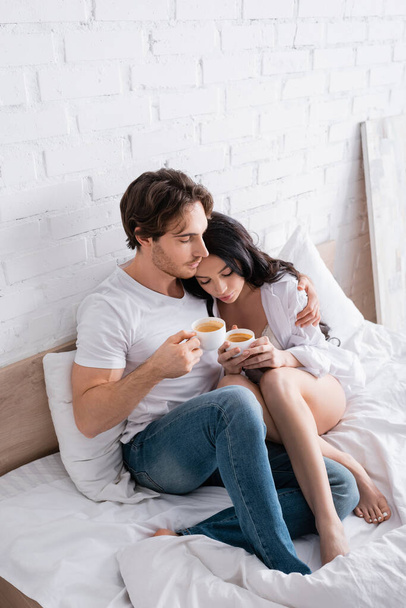 sexy young couple with cups of coffee embracing in bed with closed eyes - Photo, Image