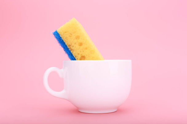 Creative, minimal dishwashing concept. Simple composition with yellow cleaning kitchen sponge in white ceramic cup on pink background  - 写真・画像