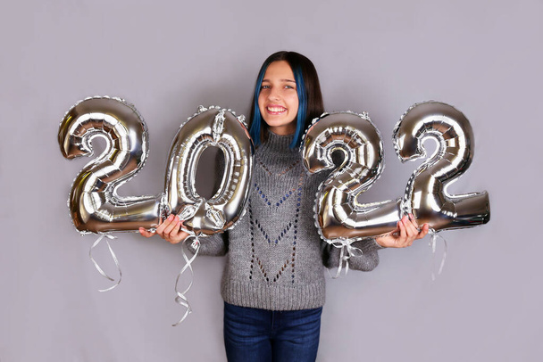 A beautiful cheerful brunette on a gray background holds silver balloons from the number 2022 in her hands. Happy New Year 2022.  - Fotografie, Obrázek