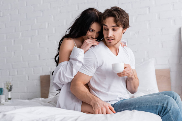 sexy brunette femme séduire jeune homme assis sur le lit avec tasse de café - Photo, image