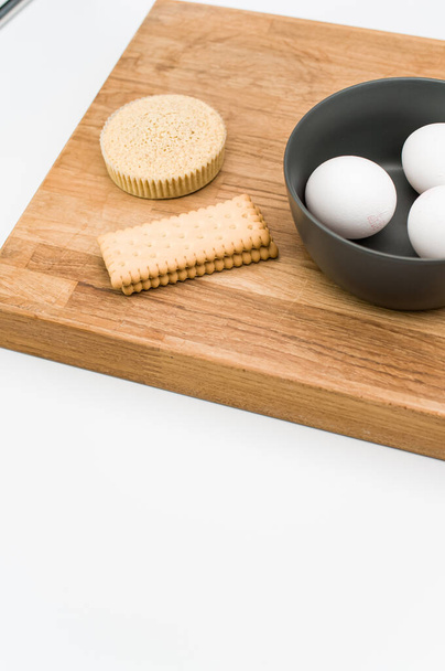 organic cake in a paper backing cup biscuits and bowl with eggs on a wooden chopping board on white background - Φωτογραφία, εικόνα