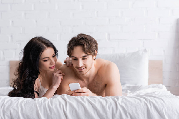 sexy hemdlosen Mann mit Smartphone in der Nähe verführerische brünette Frau im Schlafzimmer - Foto, Bild