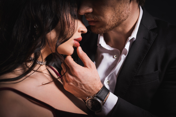 κομψός άνδρας με μαύρο κοστούμι που αγγίζει τα χείλη της αισθησιακής μελαχρινής γυναίκας  - Φωτογραφία, εικόνα