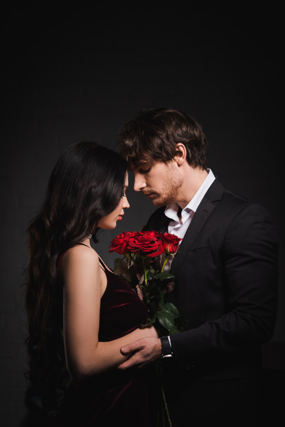 zijaanzicht van brunette vrouw met rode rozen staan oog in oog met man op donkere achtergrond - Foto, afbeelding