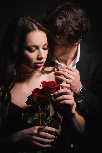 joven abrazando hombro de mujer seductora sosteniendo rosas rojas aisladas en negro - Foto, imagen